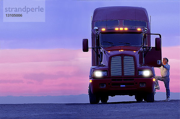 Die Silhouette eines Lkw-Fahrers  der bei Sonnenaufgang in das Fahrerhaus seiner gewerblichen Zugmaschine der Klasse 8 einsteigt.