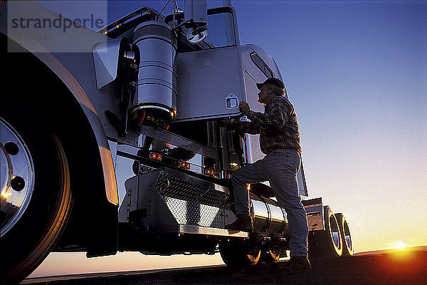 Die Silhouette eines Lkw-Fahrers  der bei Sonnenaufgang in das Fahrerhaus seiner gewerblichen Zugmaschine der Klasse 8 einsteigt.