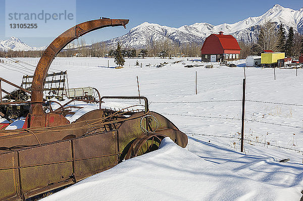 Eine Scheune mit rotem Dach steht neben alten  verrosteten landwirtschaftlichen Geräten auf einer Farm  im Hintergrund die schneebedeckten Chugach Mountains  Süd-Zentral-Alaska; Palmer  Alaska  Vereinigte Staaten von Amerika