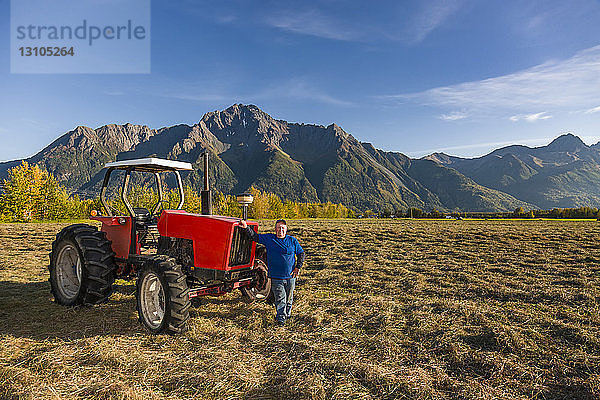 Ein Farmer in einem blauen Hemd steht neben einem alten roten Traktor auf einem offenen Feld  Pioneer Peak im Hintergrund  Süd-Zentral-Alaska; Palmer  Alaska  Vereinigte Staaten von Amerika