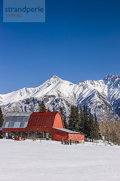 Eine Scheune mit rotem Dach in einem schneebedeckten Feld mit den Chugach Mountains im Hintergrund  Süd-Zentral-Alaska; Palmer  Alaska  Vereinigte Staaten von Amerika