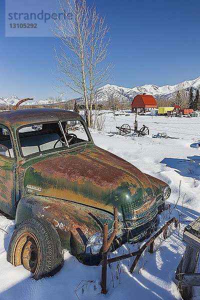 Ein alter Chevrolet-Truck und alte landwirtschaftliche Geräte  die Rost aufweisen  stehen vor einer rot überdachten Scheune auf einer Farm  im Hintergrund die schneebedeckten Chugach Mountains  Süd-Zentral-Alaska; Palmer  Alaska  Vereinigte Staaten von Amerika