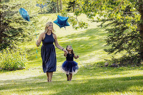 Eine Mutter  die an einem warmen Herbsttag mit ihrem kleinen Mädchen  das ein Partykleid trägt  durch einen Park spaziert  und beide tragen sternförmige Luftballons; Edmonton  Alberta  Kanada