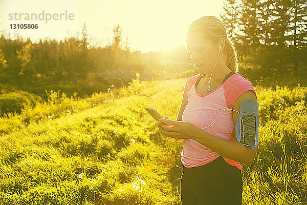 Eine attraktive Frau mittleren Alters  die Sportkleidung trägt  hält inne  um eine SMS zu schreiben  während sie an einem warmen Herbstabend in einem Stadtpark neben einem Bach läuft und Musik auf ihrem Smartphone hört; Edmonton  Alberta  Kanada