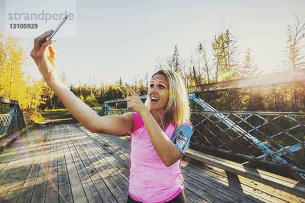 Eine Frau  die Sportkleidung und ein Armband für ihr Mobiltelefon trägt  steht im Herbst auf einer Brücke in einem Park und macht ein Selbstporträt mit ihrem Mobiltelefon; Edmonton  Alberta  Kanada