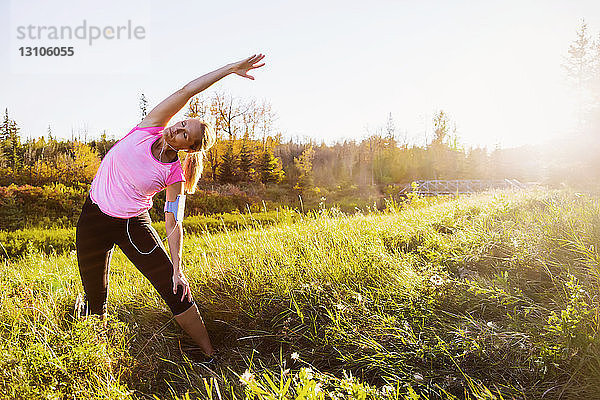 Eine attraktive Frau mittleren Alters trägt Sportkleidung und hört Musik  während sie während eines Laufs in einem Stadtpark bei Sonnenuntergang an einem warmen Herbstabend einige Dehnübungen macht; Edmonton  Alberta  Kanada