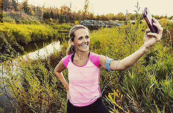Eine attraktive Frau mittleren Alters  die Sportkleidung trägt  hält inne  um ein Selbstporträt aufzunehmen  während sie an einem warmen Herbstabend an einem Bach in einem Stadtpark läuft und dabei Musik auf ihrem Smartphone hört; Edmonton  Alberta  Kanada