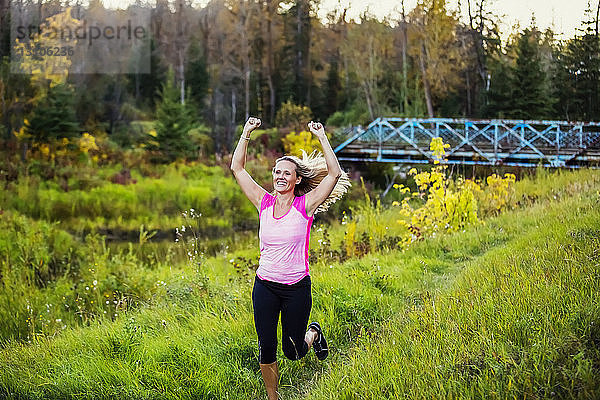 Eine attraktive Frau mittleren Alters in Sportkleidung  die an einem warmen Herbstabend bei Sonnenuntergang in einem Stadtpark an einem Bach entlang läuft und dabei die Hände in die Luft streckt; Edmonton  Alberta  Kanada