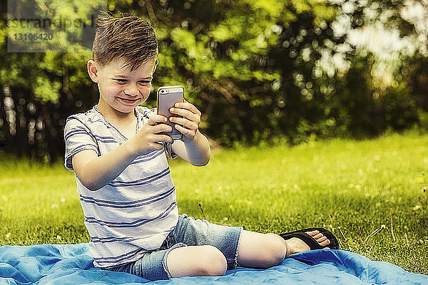 Ein kleiner Junge macht alberne Gesichtsausdrücke  während er an einem warmen Sommertag in einem Stadtpark Selbstporträts mit einem Smartphone aufnimmt; Edmonton  Alberta  Kanada