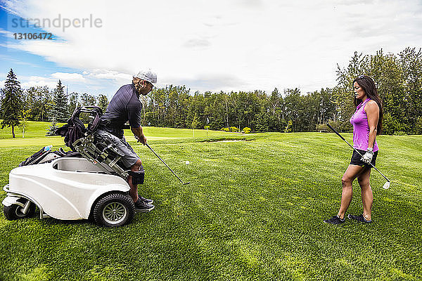 Ein körperlich behinderter Golfer  der einen Spezialrollstuhl benutzt  schlägt den Golfball mit seinem Golfschläger auf dem Golfplatz  während eine Golferin zusieht; Edmonton  Alberta  Kanada
