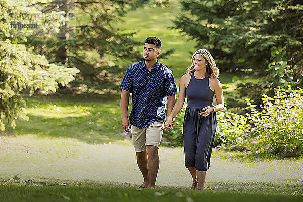 Ein gemischtrassiges Ehepaar geht in einem Stadtpark spazieren und hält sich an den Händen  während sie an einem warmen  sonnigen Nachmittag Zeit miteinander verbringen; Edmonton  Alberta  Kanada