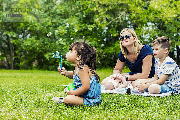 Eine junge Mutter beobachtet ihre Tochter beim Blasen von Seifenblasen  während sie mit ihrem Baby und ihrem Sohn auf einer Decke in einem Stadtpark an einem warmen sonnigen Tag sitzt; Edmonton  Alberta  Kanada