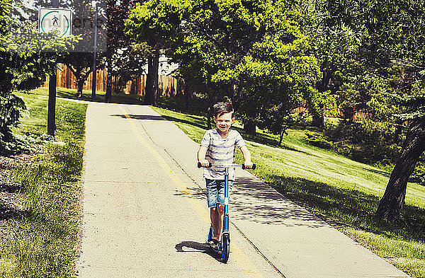 Ein kleiner Junge fährt mit seinem Roller auf einem Weg in einem Stadtpark und lächelt in die Kamera: Edmonton  Alberta  Kanada