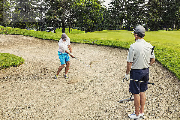 Zwei männliche Golfer stehen in einem Bunker  einer hält eine Harke und wartet  bis er an der Reihe ist  während der andere den Ball mit einem Chip aus der Sandgrube herausholt; Edmonton  Alberta  Kanada