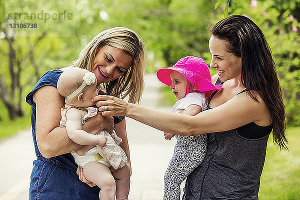 Zwei junge Mütter halten inne  während sie an einem warmen Sommertag mit ihren kleinen Mädchen in einem Stadtpark spazieren gehen  die neugierig aufeinander sind; Edmonton  Alberta  Kanada