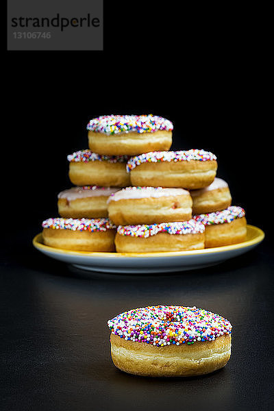 Nahaufnahme eines mit Süßigkeiten bedeckten Donuts mit einem Teller voller mit Süßigkeiten bedeckter Donuts im Hintergrund auf einem schwarzen reflektierenden Hintergrund