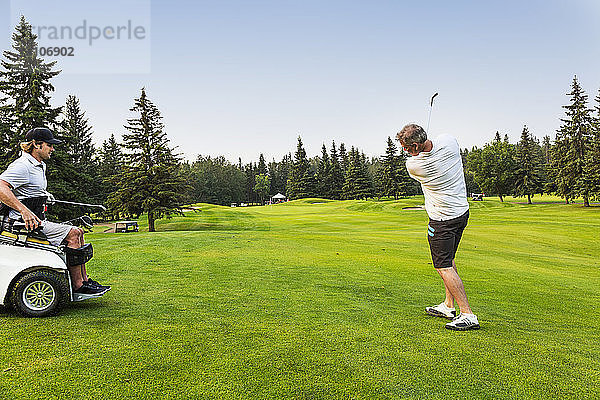 Ein männlicher Golfer schlägt einen Golfball mit einem Wedge auf einem Golfplatz auf das Grün  während ein behinderter Golfer in einem Spezialrollstuhl zusieht; Edmonton  Alberta  Kanada