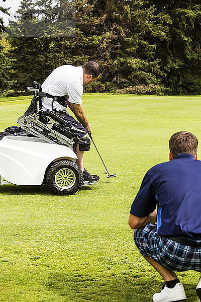 Ein nichtbehinderter Golfer hilft einem behinderten Golfer  der einen speziellen elektrischen Golfrollstuhl benutzt  seinen Putt auf dem Grün eines Golfplatzes zu platzieren; Edmonton  Alberta  Kanada