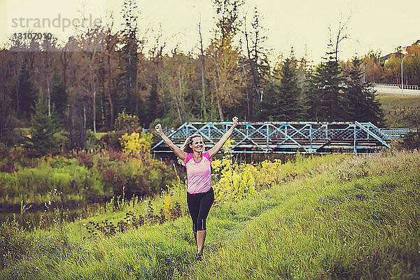 Eine attraktive Frau mittleren Alters in Sportkleidung  die an einem warmen Herbstabend bei Sonnenuntergang in einem Stadtpark an einem Bach entlang läuft und dabei die Hände in die Luft streckt; Edmonton  Alberta  Kanada