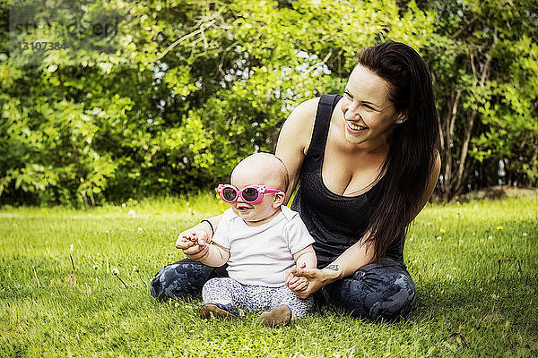 Porträt einer jungen Mutter  die im Sommer Zeit mit ihrer Tochter in einem Park verbringt  wobei das Baby eine lustige Sonnenbrille trägt; Edmonton  Alberta  Kanada