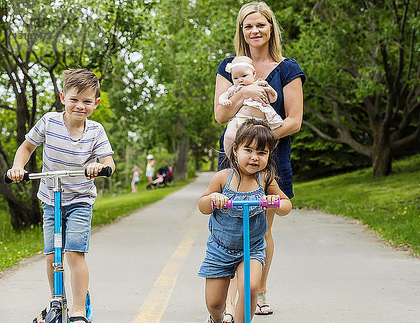 Eine junge Mutter geht spazieren und hält ihr Baby  während sie ihren anderen Kindern beim Rollerfahren auf einem Weg in einem Park an einem warmen Sommertag zusieht; Edmonton  Alberta  Kanada