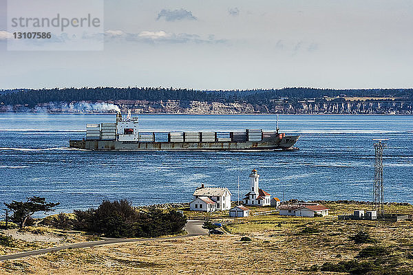 Frachtschiff  das von der Straße von Juan de Fuca über Admiralty Inlet in den Puget Sound einfährt. Der Point Wilson-Leuchtturm befindet sich im Fort Worden Historical State Park in der Nähe der Stadt Port Townsend. Im Hintergrund ist Whidbey Island zu sehen; Washington  Vereinigte Staaten von Amerika