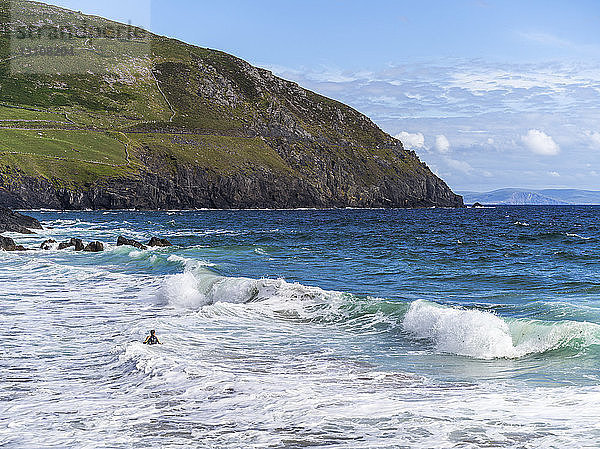 Zerklüftete Küstenlinie mit Klippen und Bergen und einem Schwimmer in den Meereswellen im Vordergrund; Ballyferriter  County Kerry  Irland