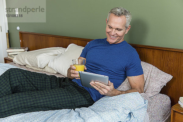 Lächelnder reifer Mann  der Saft hält  während er zu Hause am Bett einen Tablettencomputer benutzt