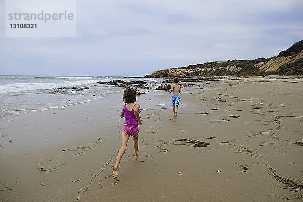 Rückansicht der Geschwister  die im Crystal Cove State Park am Strand gegen den Himmel rennen