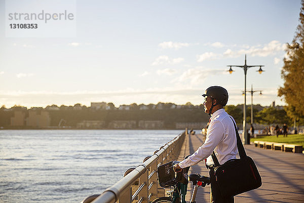 Geschäftsmann mit Fahrrad schaut weg  während er bei Sonnenuntergang am Fluss steht