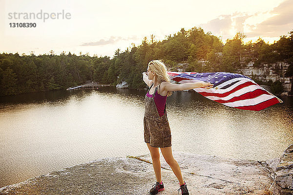 Junge Frau hält amerikanische Flagge  während sie auf einem Felsen am See steht