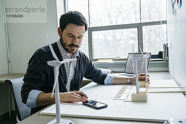 Geschäftsmann benutzt Smartphone  während er Modelle von Windkraftanlagen auf dem Schreibtisch arrangiert