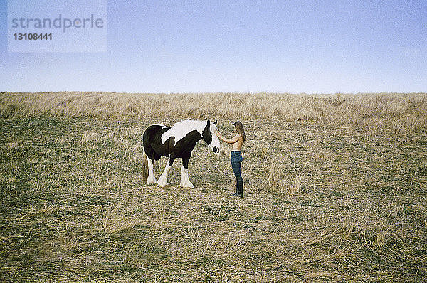 Seitenansicht einer Oben-ohne-Frau  die ihr Pferd auf einem Grasfeld vor klarem Himmel streichelt