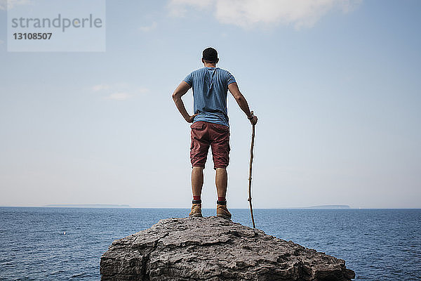 Rückansicht eines Wanderers mit der Hand an der Hüfte  der einen Stock hält  während er auf einem Felsen gegen Meer und Himmel im Bruce Peninsula National Park steht