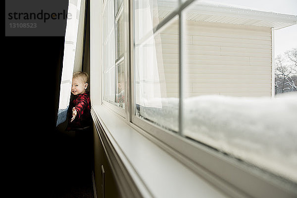 Porträt eines verspielten Jungen  der zu Hause hinter Vorhängen steht