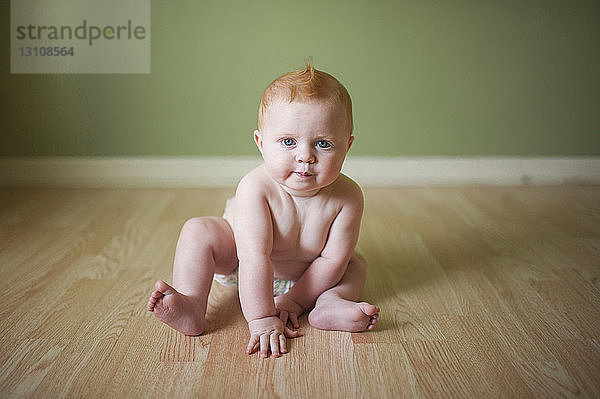 Porträt eines niedlichen Jungen ohne Hemd  der zu Hause auf dem Boden an der Wand sitzt