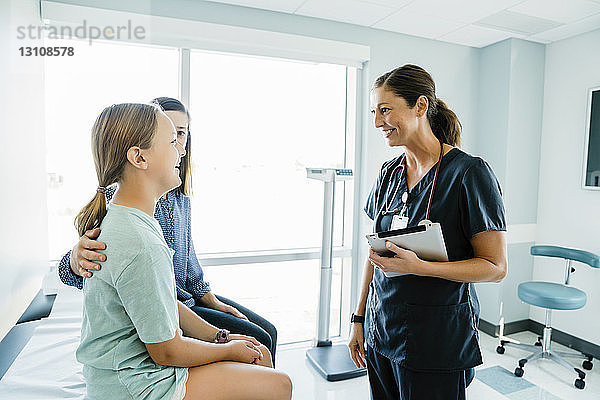 Fröhliches Mädchen schaut den Kinderarzt an  während sie im Krankenhaus neben der Mutter auf dem Untersuchungstisch sitzt