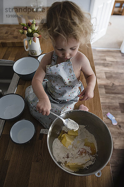 Schrägansicht eines Mädchens  das Teig in einer Schüssel mischt  während es auf der Küchentheke sitzt