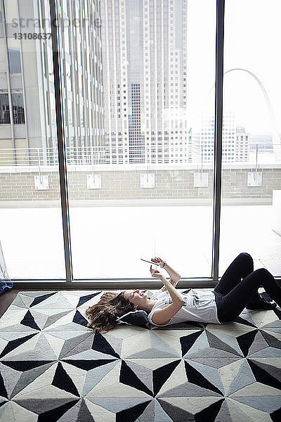 Frau  die ein Smartphone mit hohem Blickwinkel benutzt  während sie zu Hause am Fenster auf dem Teppich liegt