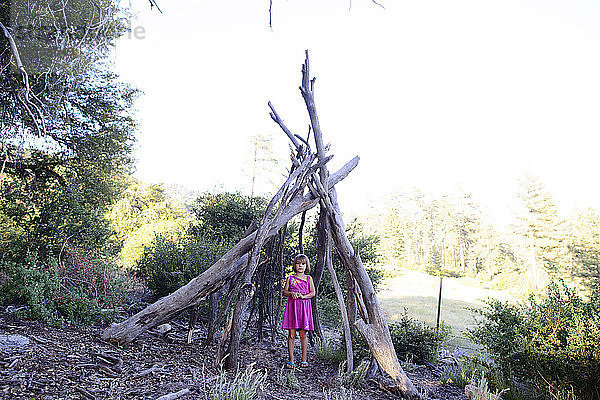 Mädchen in voller Länge steht unter einem Holzzelt im Wald vor klarem Himmel während eines sonnigen Tages