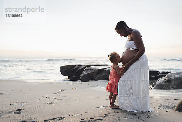 Tochter küsst schwangeren Mutterbauch  während sie bei Sonnenuntergang am Strand steht