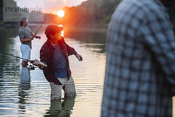 Mittelteil eines älteren Mannes  der im See steht  während Freunde bei Sonnenuntergang im Hintergrund fischen