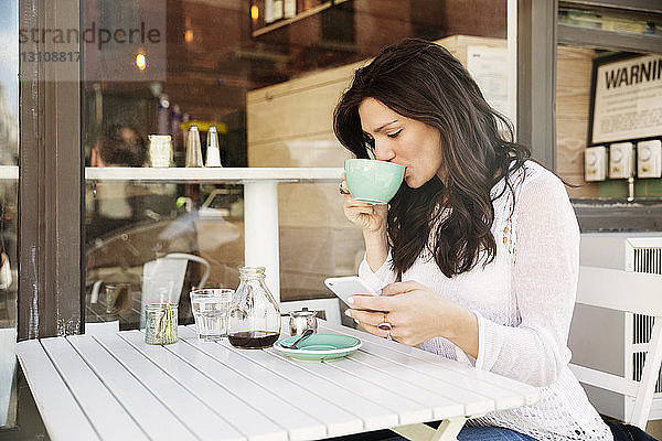 Frau trinkt Kaffee  während sie im Straßencafé ein Smartphone benutzt