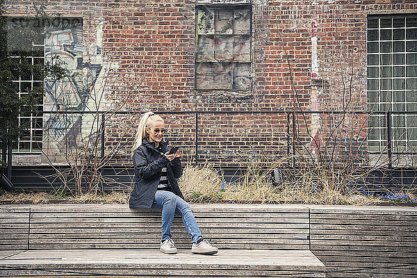 Eine Frau in voller Länge benutzt ein Mobiltelefon  während sie auf einer Stützmauer gegen ein Gebäude in der Stadt sitzt