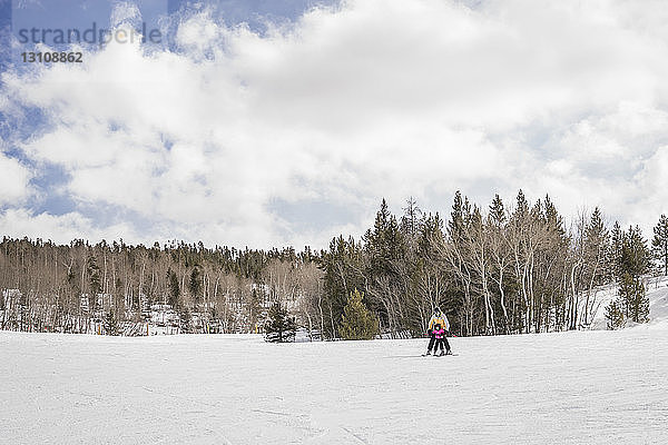 Vater mit Tochter beim Skifahren auf schneebedeckter Landschaft vor bewölktem Himmel im Wald