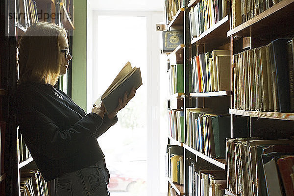 Seitenansicht einer Frau  die ein Buch liest  während sie sich an ein Bücherregal lehnt