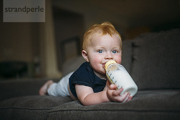 Porträt eines süßen kleinen Jungen  der Milch aus der Flasche trinkt  während er zu Hause auf dem Sofa liegt