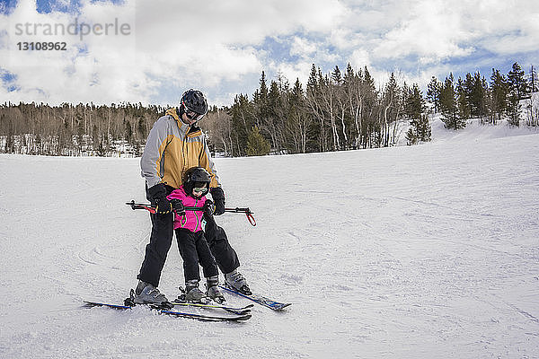 Vater mit Tochter beim Skifahren in schneebedeckter Landschaft vor bewölktem Himmel im Wald