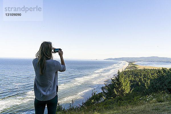 Rückansicht einer Frau  die das Meer fotografiert  während sie gegen den klaren Himmel steht