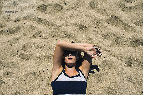 Draufsicht einer Frau  die ihr Gesicht bedeckt  während sie am sonnigen Tag auf Sand liegt